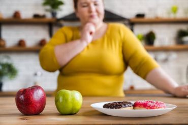 A larger woman deciding whether to eat the red or green apples or the iced donuts on the table in front of her.