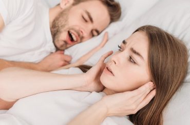 A young couple lying in bed. The male is snoring loudly and his partner is holding her hands over her ears to block the snoring sound.