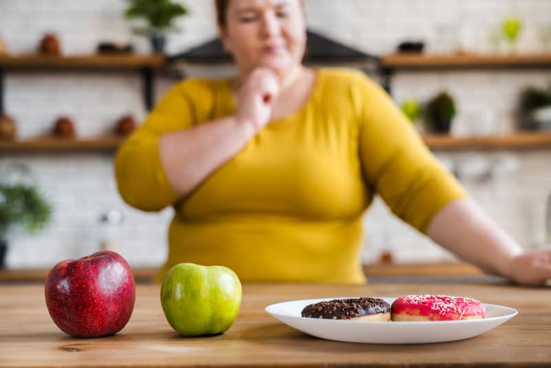 A larger woman deciding whether to eat the red or green apples or the iced donuts on the table in front of her.